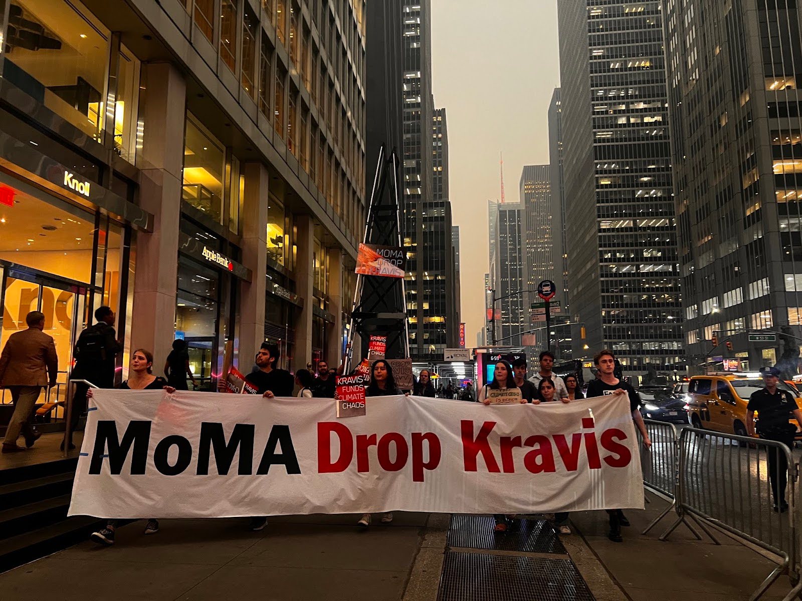 A crowd of activists marching in a city street holding the banner "MoMA Drop Kravis"